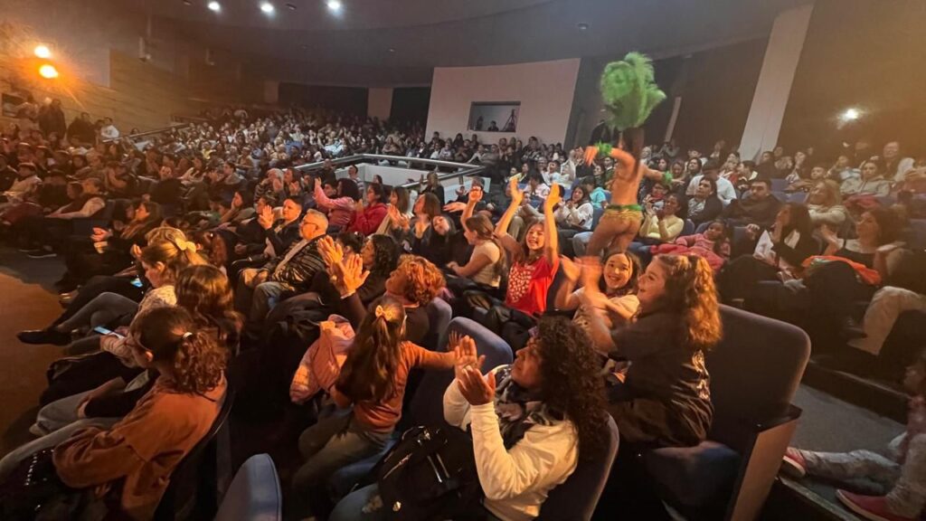 Imagen de gente en la platea de un teatro aplaudiendo mientras se observan pasantes de comparsa por entre el público.