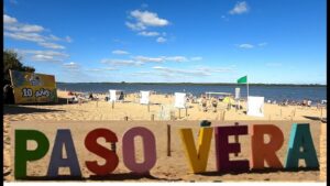 Cartel Paso Vera de coleres sobre la arena y de fondo rìo y vegetaciòn con gente bajo el sol